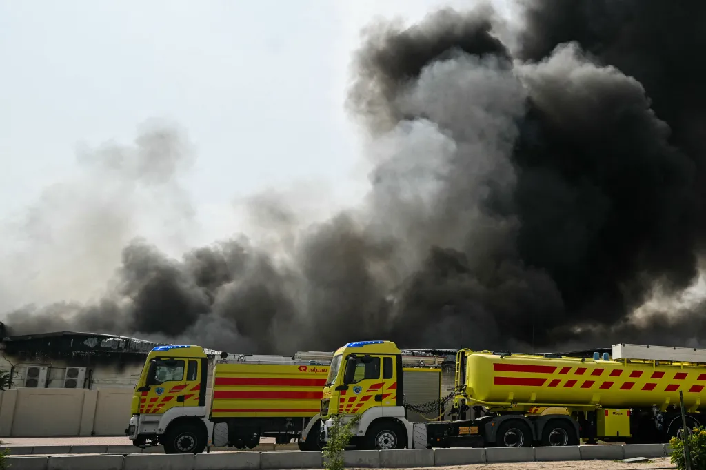 Two yellow fire trucks parked in front of a building engulfed in a large plume of black smoke.