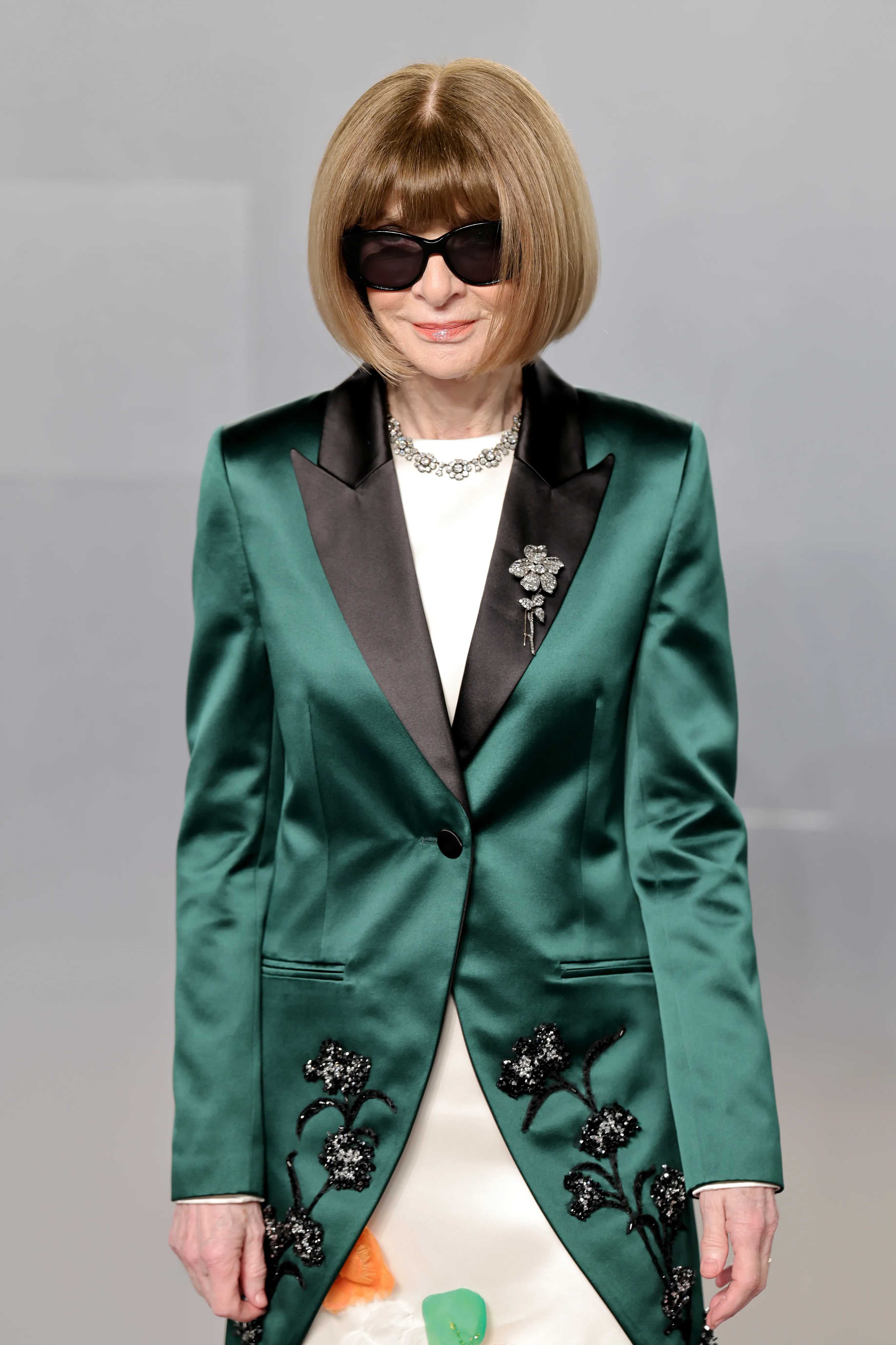Anna Wintour wearing a green blazer with black lapels and black floral embellishments, a white dress, a jeweled necklace, and sunglasses.