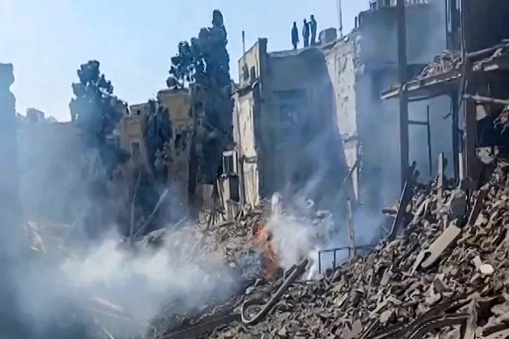 Damage at an impact site following US and Israeli strikes on Tehran, with smoke and fire amid rubble, and people inspecting the damage on a distant rooftop.