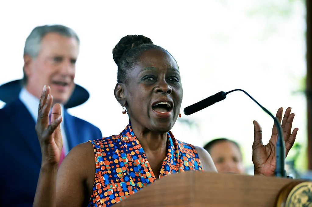 A woman in a blue, orange, and white patterned top speaks passionately into a microphone at a podium, gesturing with her hands.