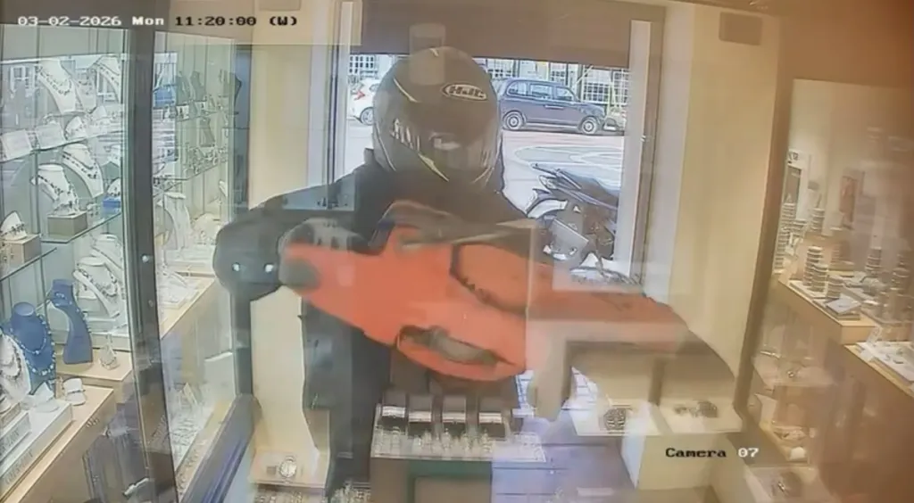 A masked person in a helmet uses a chainsaw to break into a jewelry store, with displays of necklaces and rings visible.