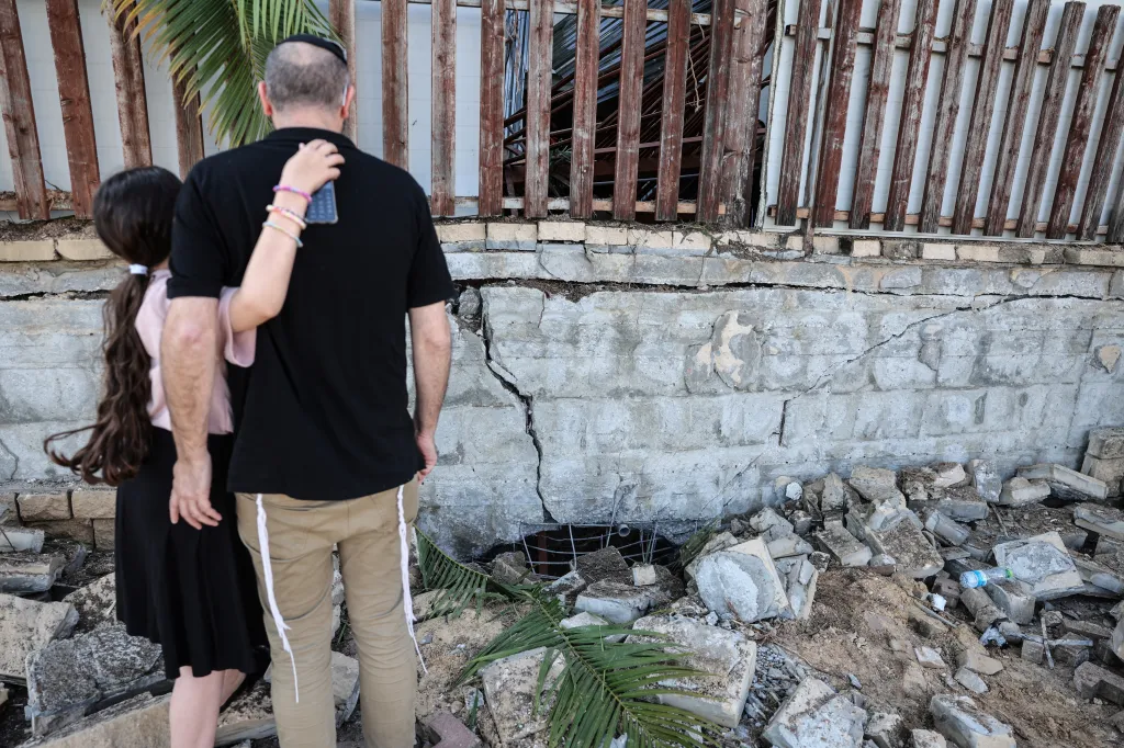 A man and a girl stand in front of a damaged building, with rubble and cracks in the wall.