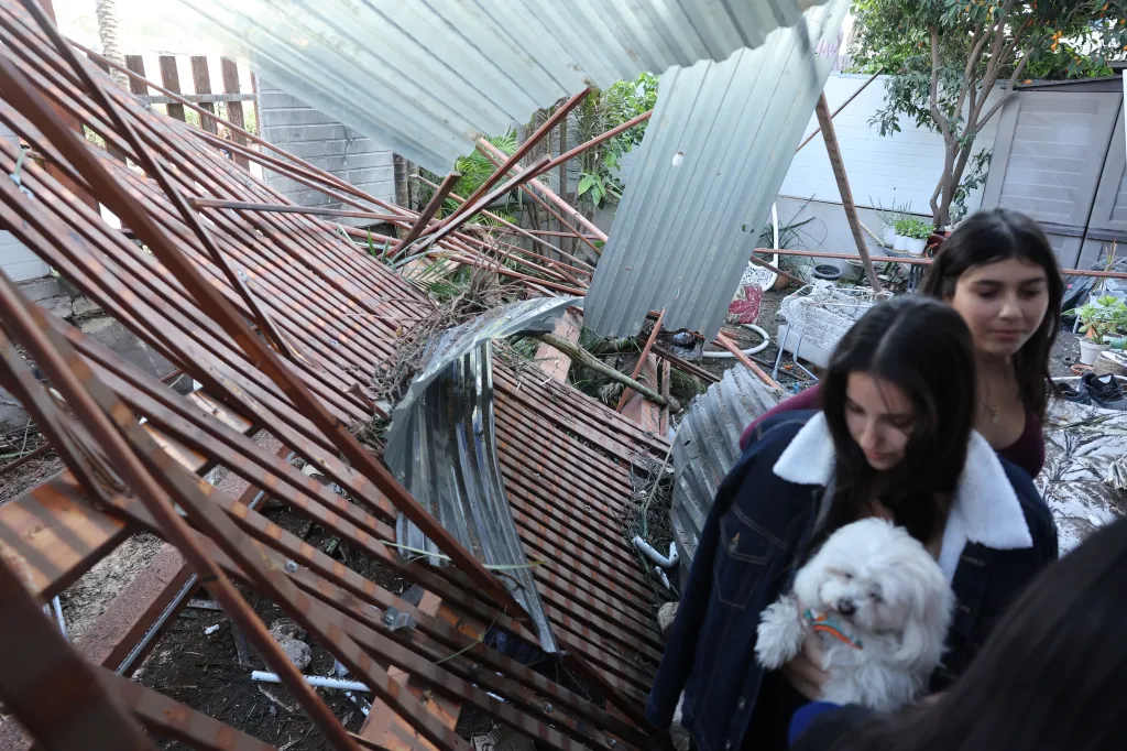 Two women with a dog standing amidst damaged corrugated metal and fallen debris.