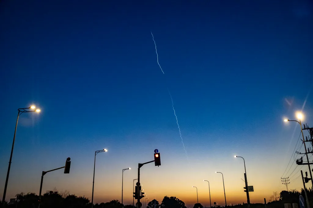 A missile contrail visible in the sky above a street at dusk.