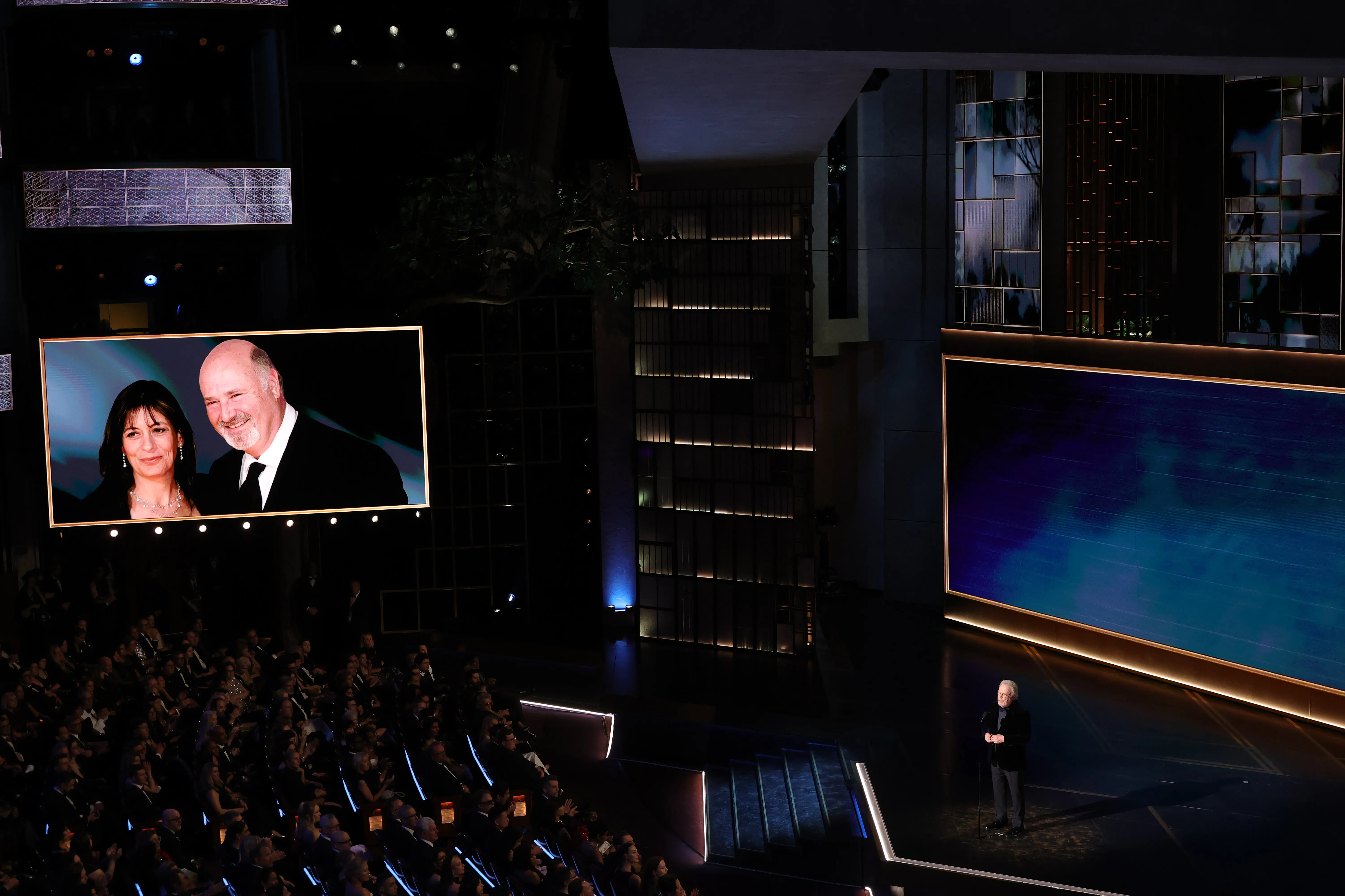 A large audience looks on as a man stands on stage at an awards show, and a screen behind him displays a picture of a man and a woman.