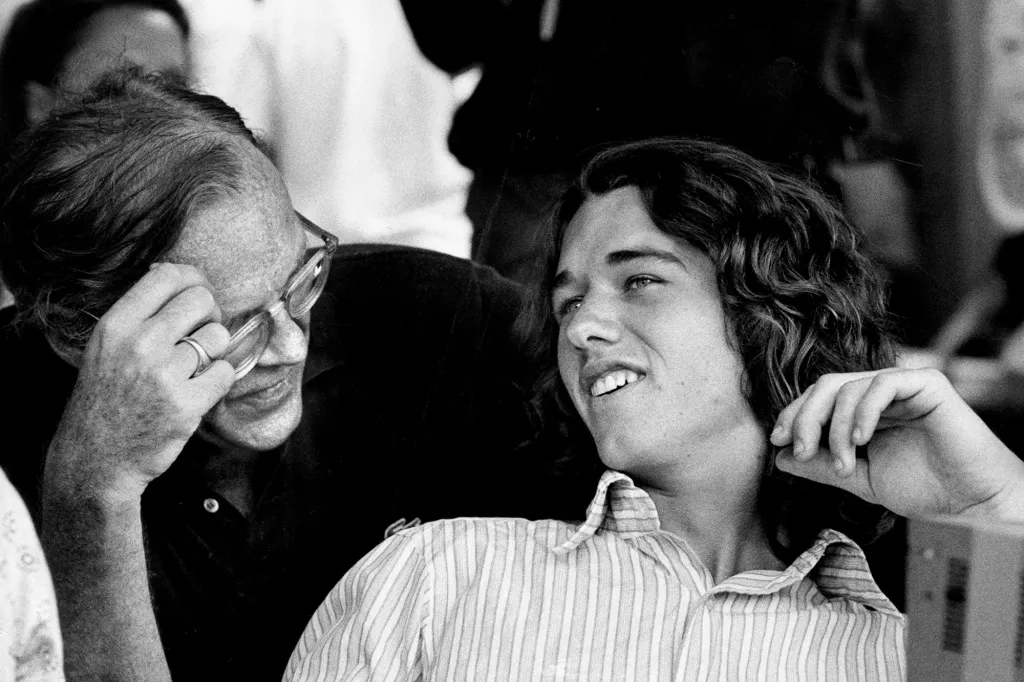 Two men, one older with glasses and a younger man with long hair, are looking at something to the right of the frame.