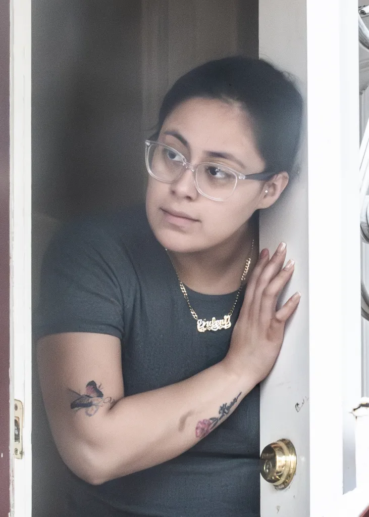 A young woman in clear glasses and a gold necklace peeks out from a doorway.