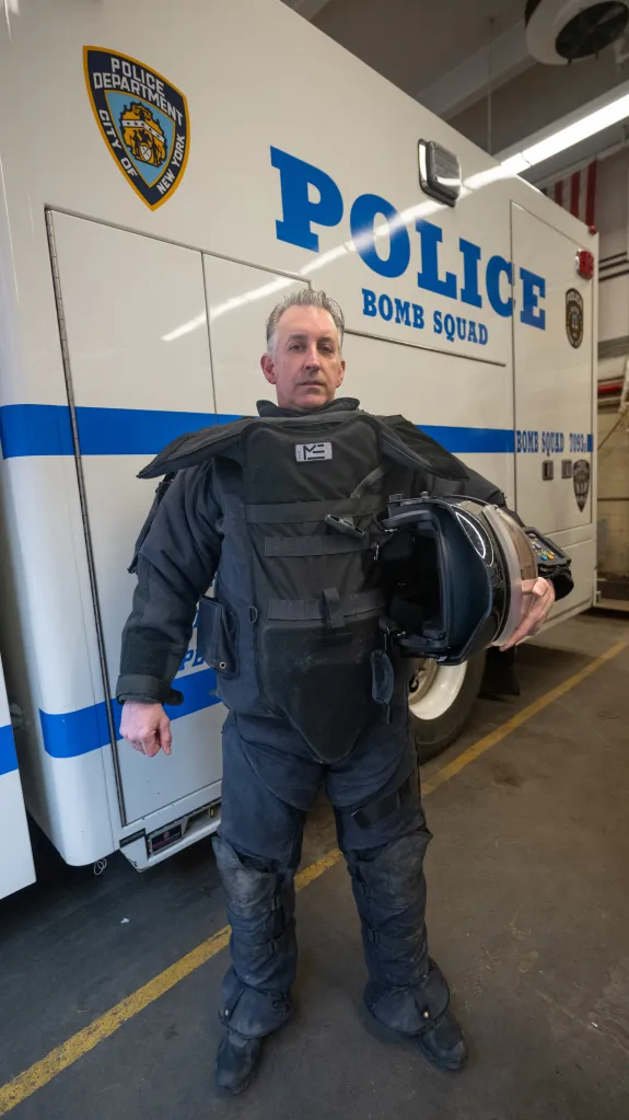Detective Sean Mulcahy in bomb squad suit, holding a helmet, standing in front of a NYPD bomb squad vehicle.