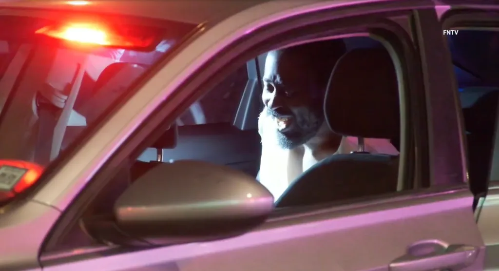 A man with a beard yelling from the driver's seat of a car, with a flashing red light visible in the reflection of the car's window.