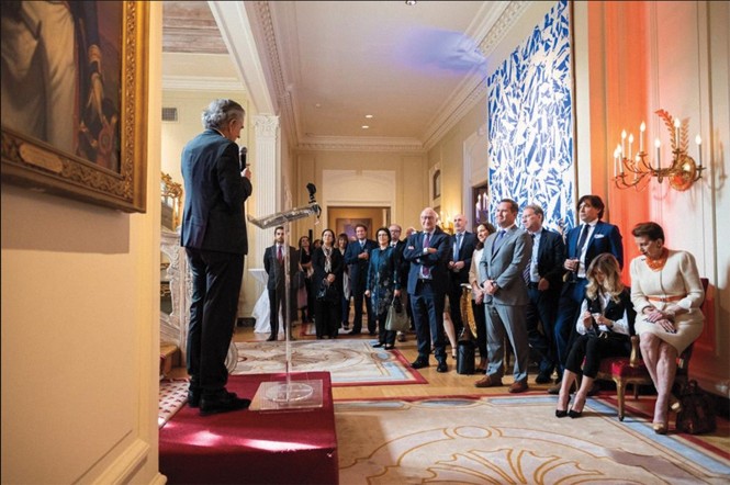 photo of embassy interior with person giving speech on slightly raised platform and small group of guests gathered watching