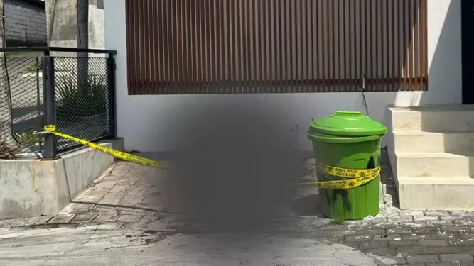 A crime scene with yellow tape wrapped around a black fence and a green trash can, obscuring the middle of the image.