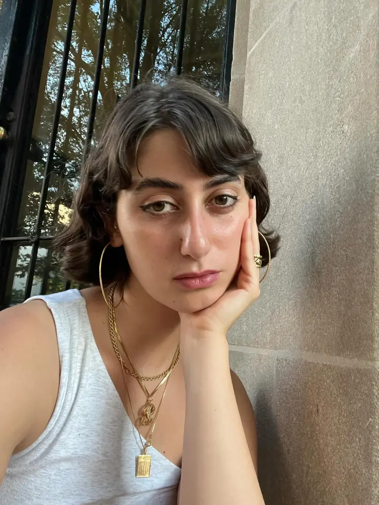 A woman with short brown hair, wearing a white tank top and multiple gold necklaces, leans her face on her hand.