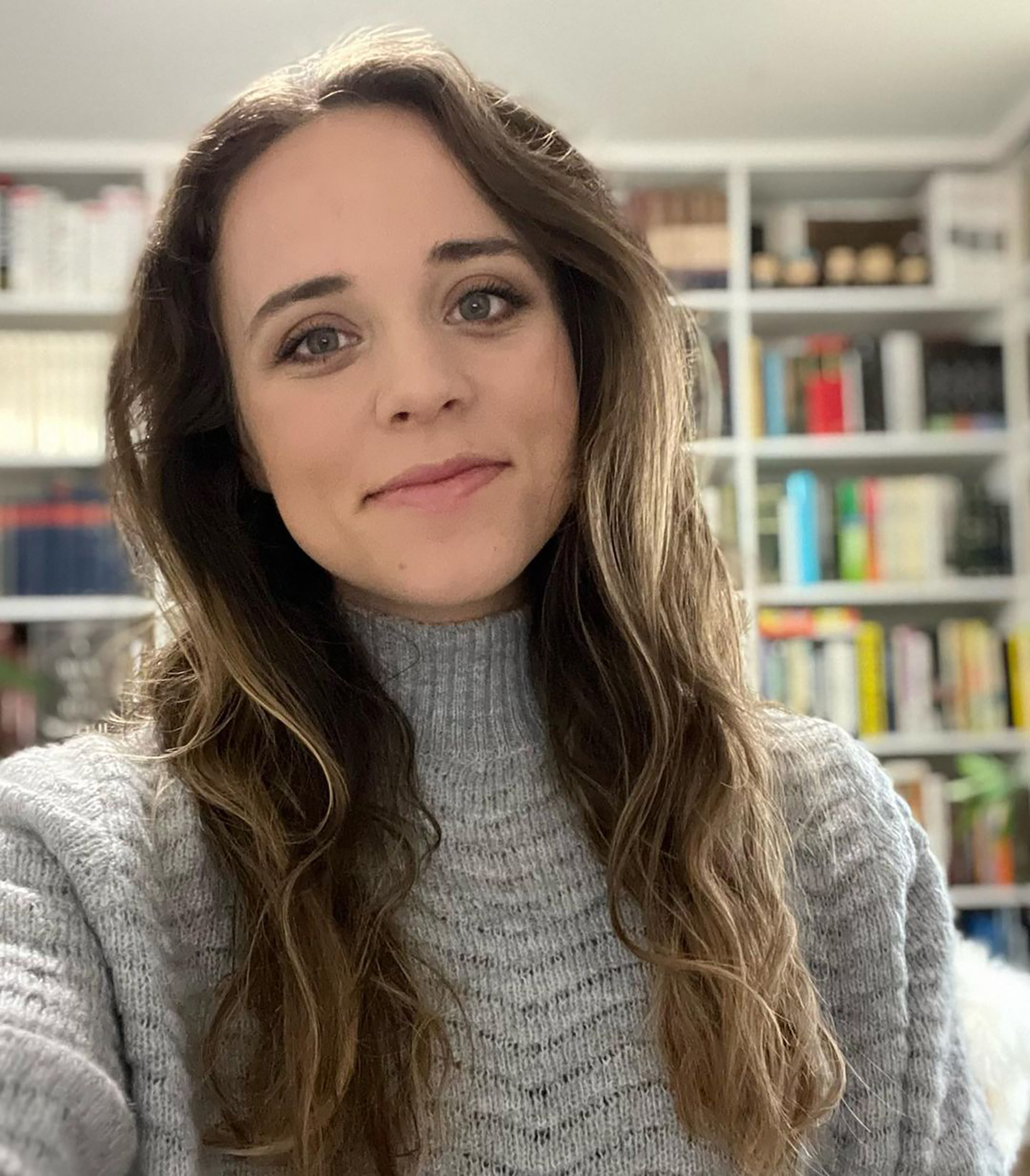 A woman with long wavy brown hair wearing a gray knitted sweater smiles at the camera, with a bookshelf visible behind her.
