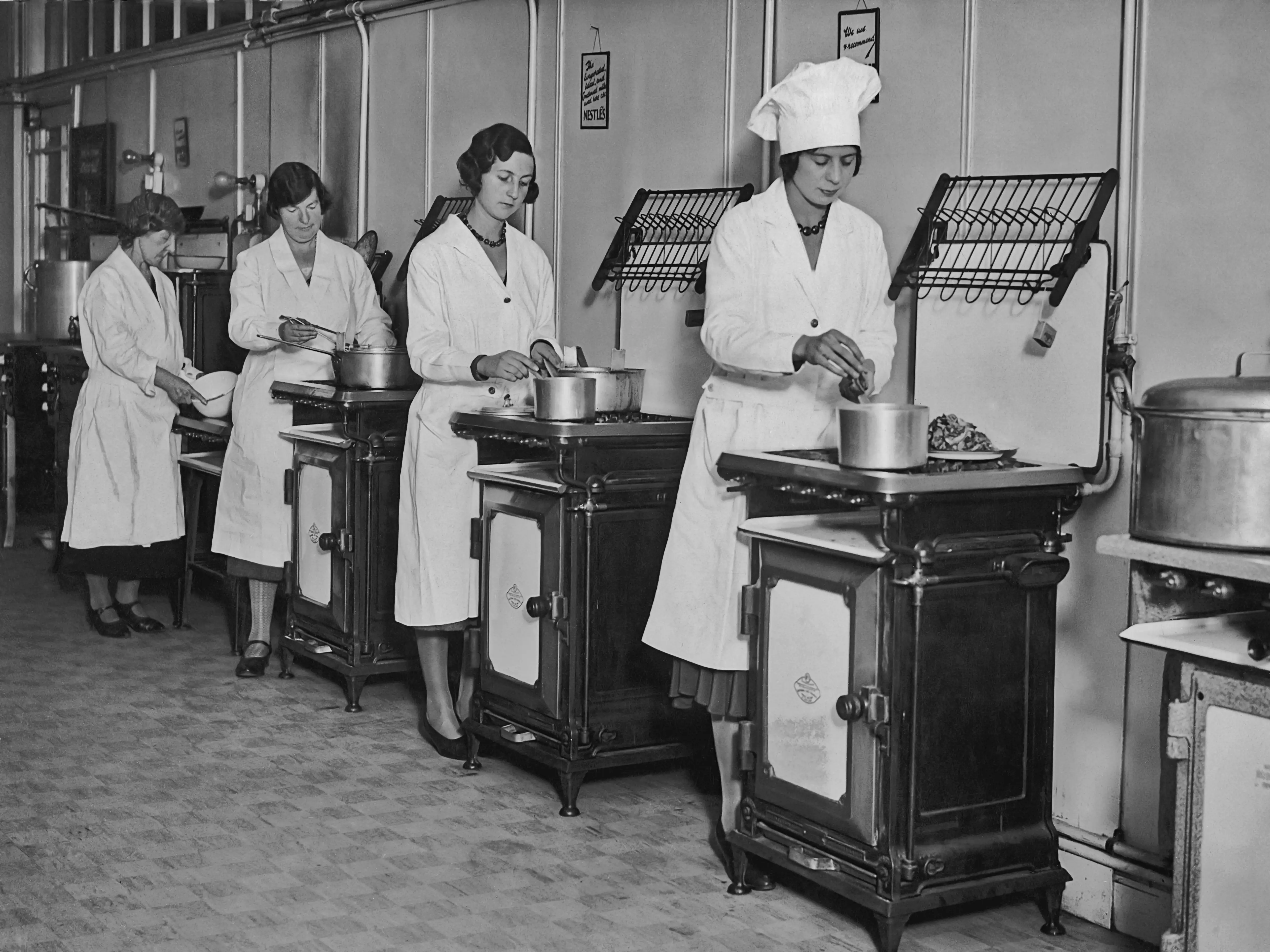 Women taking a cooking class in the 1920s; each of them stood by their own stove and wore white coats over their dresses.