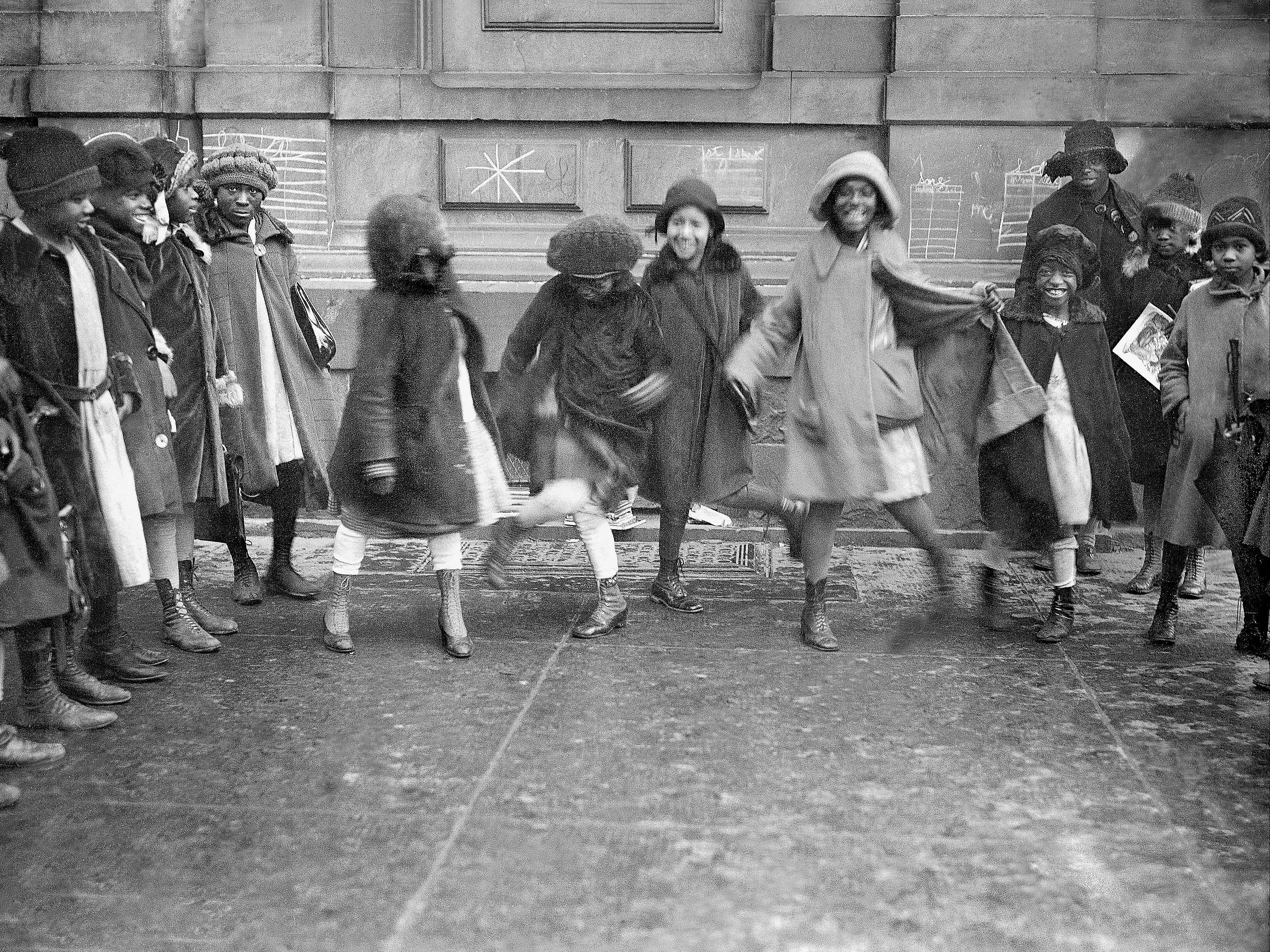 A group of Black girls smiling and dancing in Harlem, New York City, in 1920. They wore coats and hats.