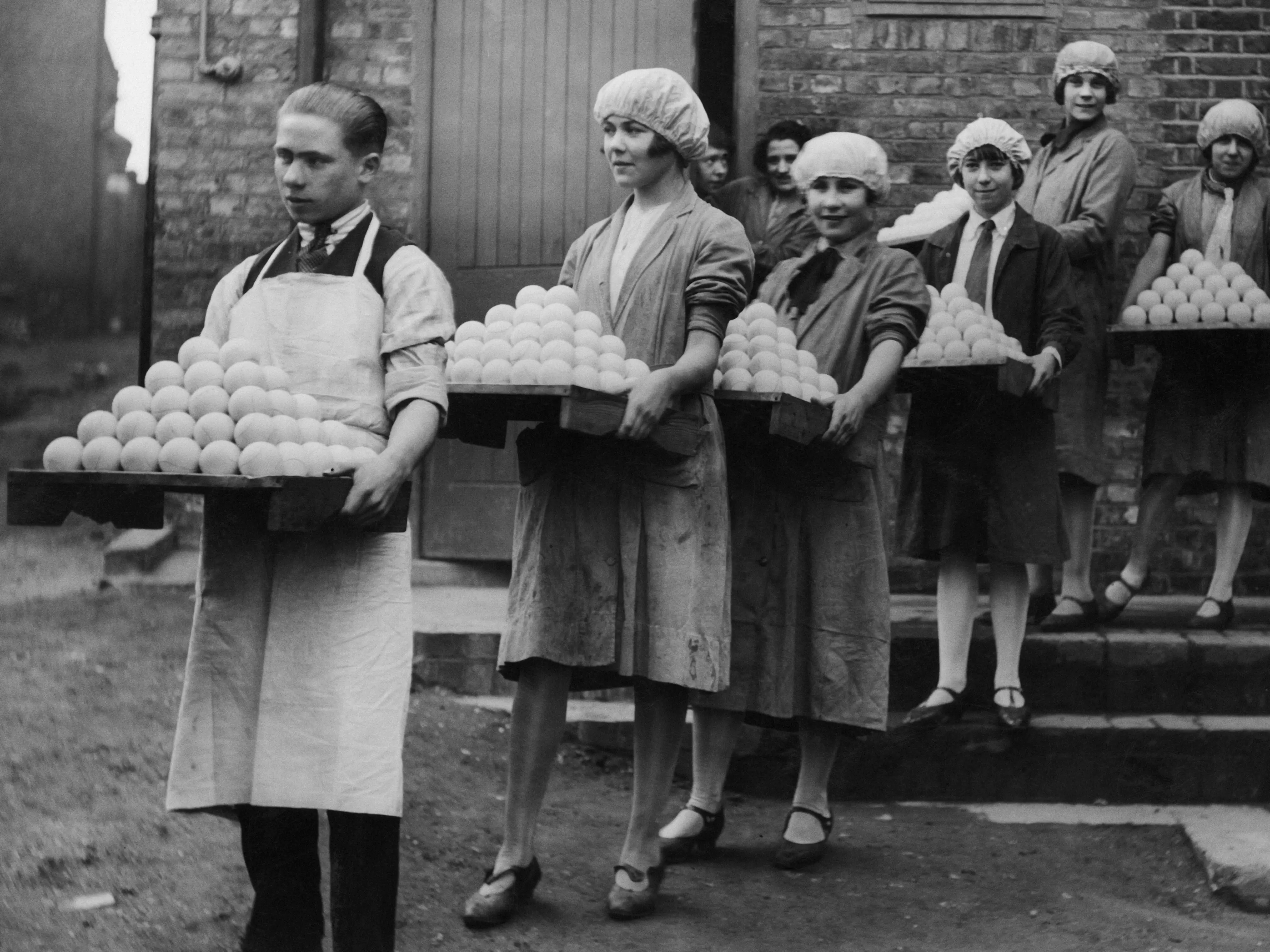 Workers, one man and five women, carrying trays of freshly manufactured tennis balls.