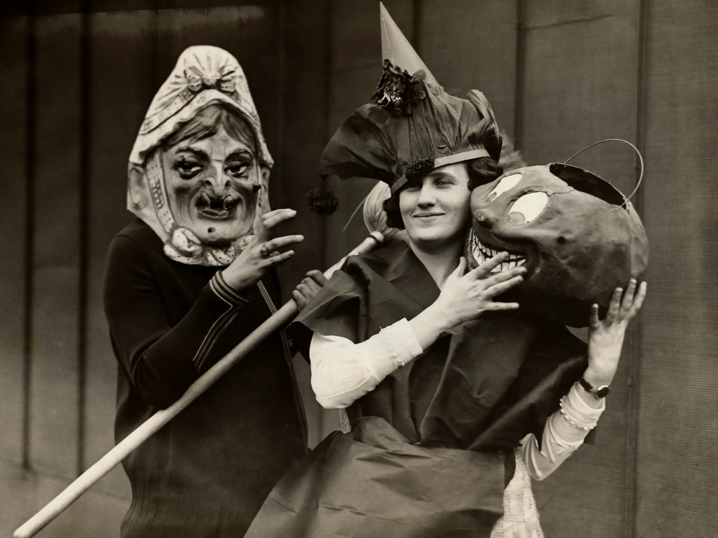 Women in the 1920s dressed up for Halloween; one is wearing a witch mask and carrying a broom while the other appears to hold a basket for candy that resembles a cartoon head.