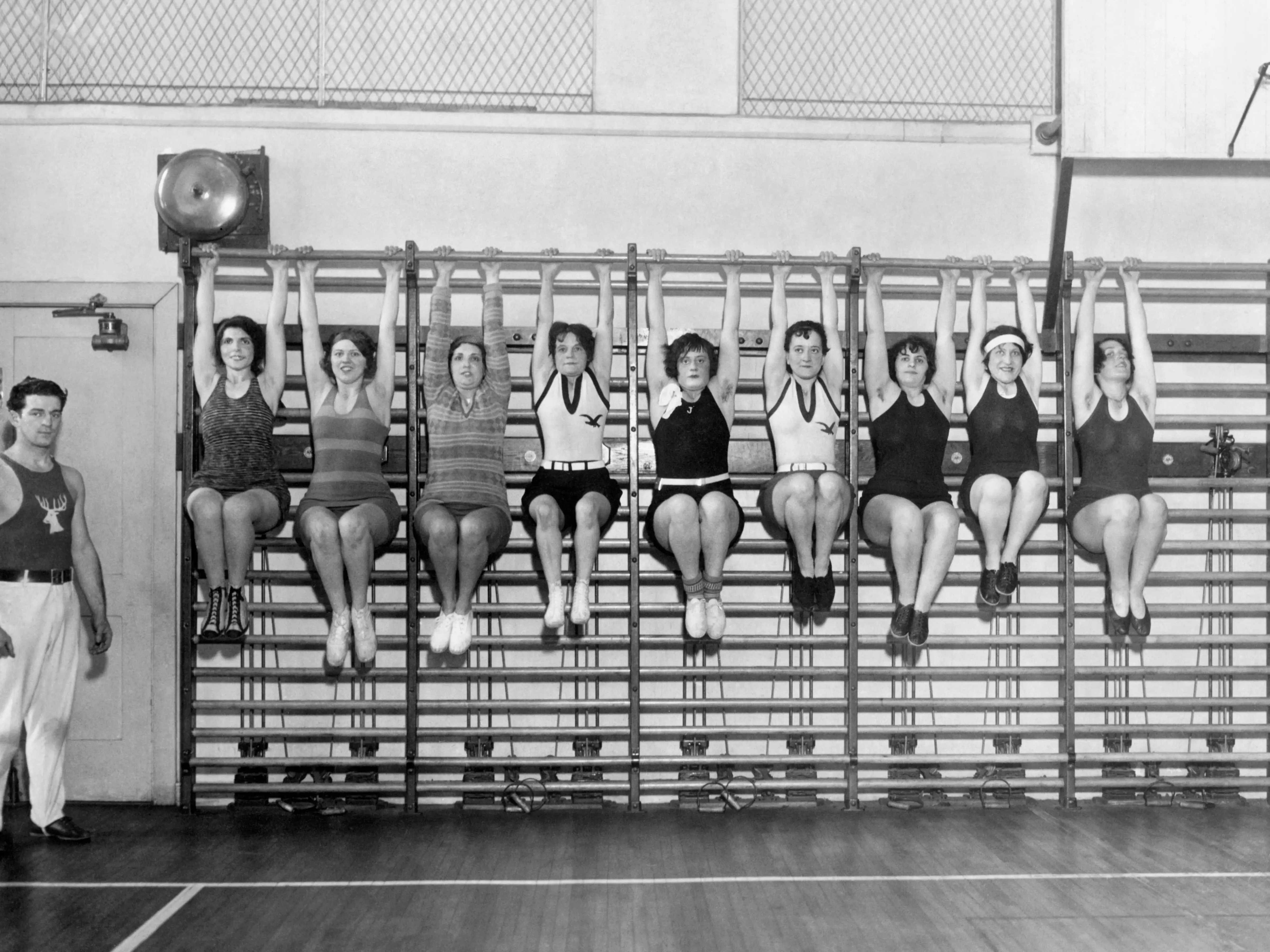 Wives of the members of the Philadelphia Elks organization working out. The nine women all hung from a bar with their knees raised at a 90 degree angle, while a man stood nearby.