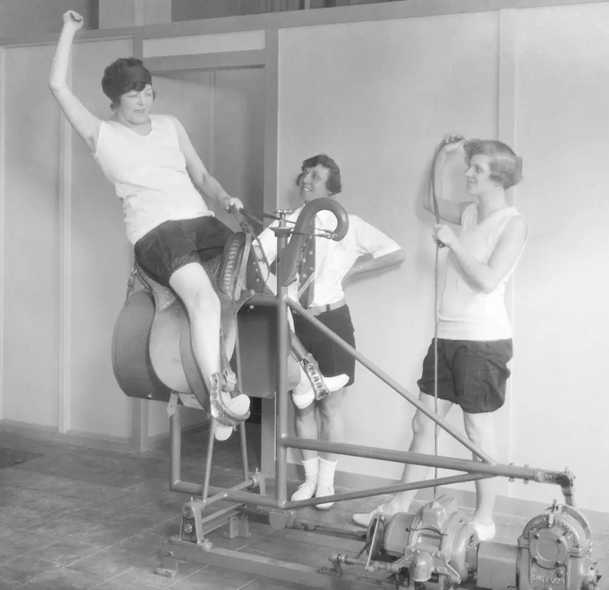A woman riding a 1920s mechanical bull while two women stood nearby and watched.