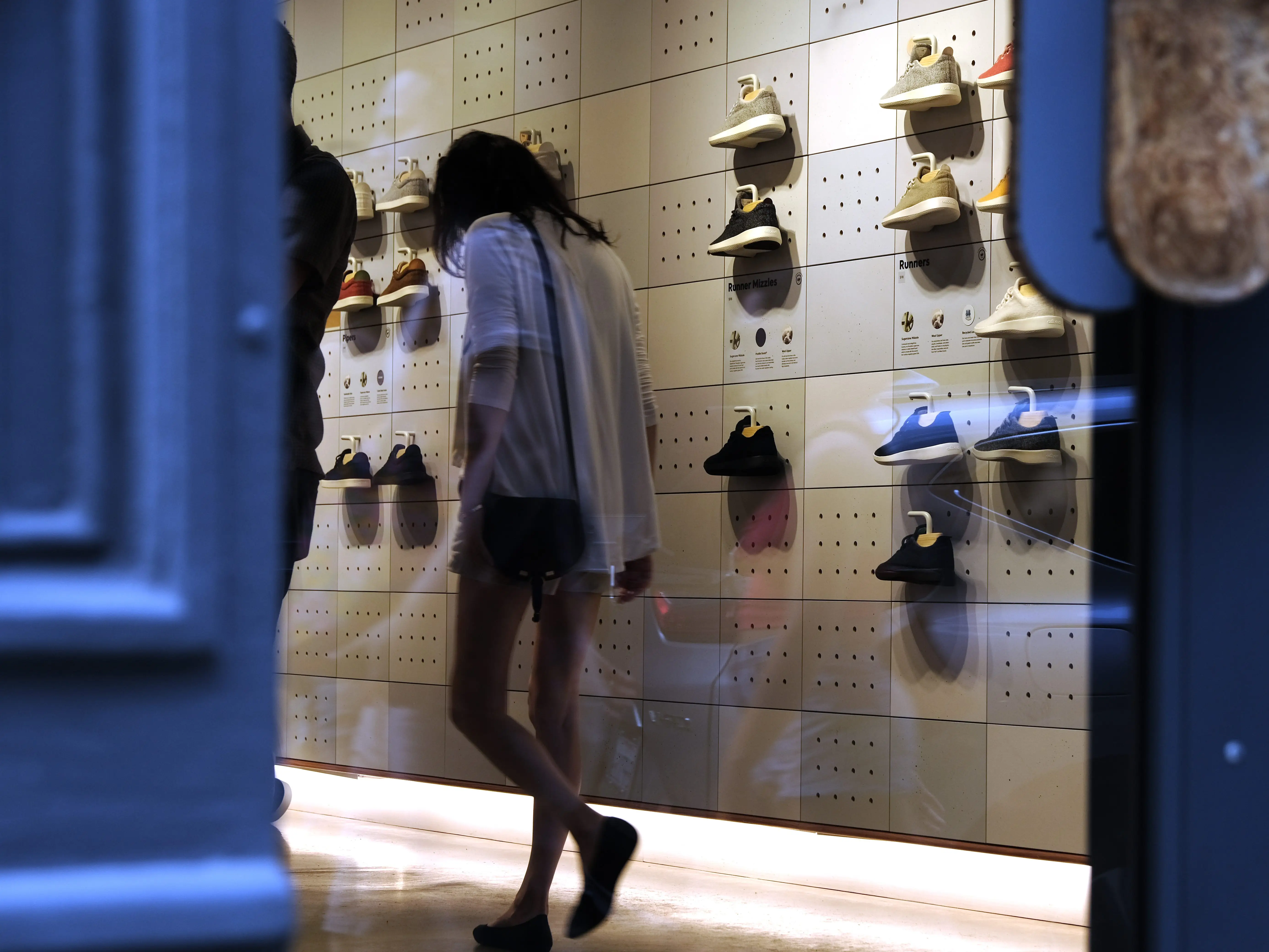 A woman walking inside an Allbirds store, with sneakers on the wall behind her.
