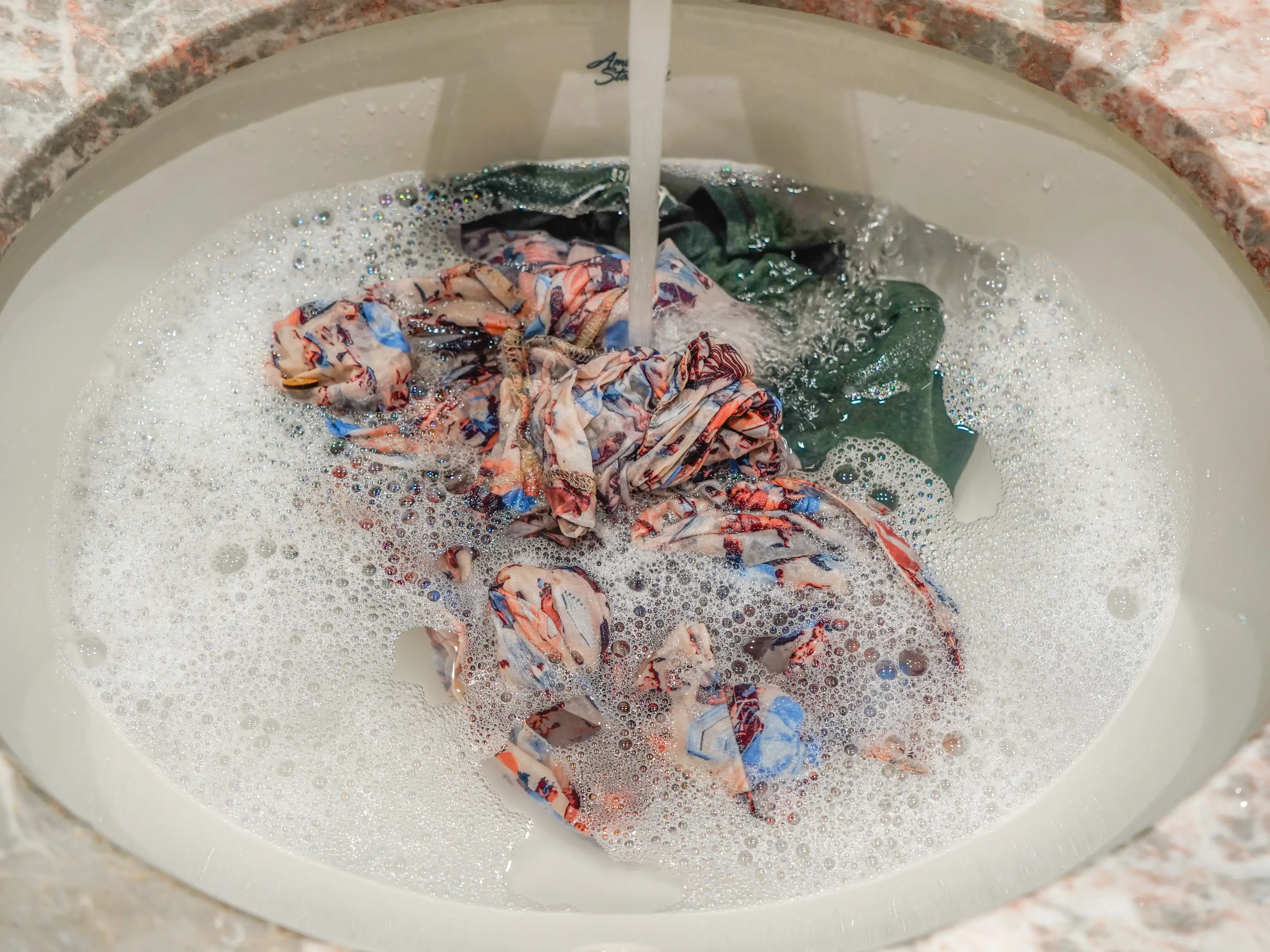 Green and floral clothing being washed with soapy water in a bathroom sink.