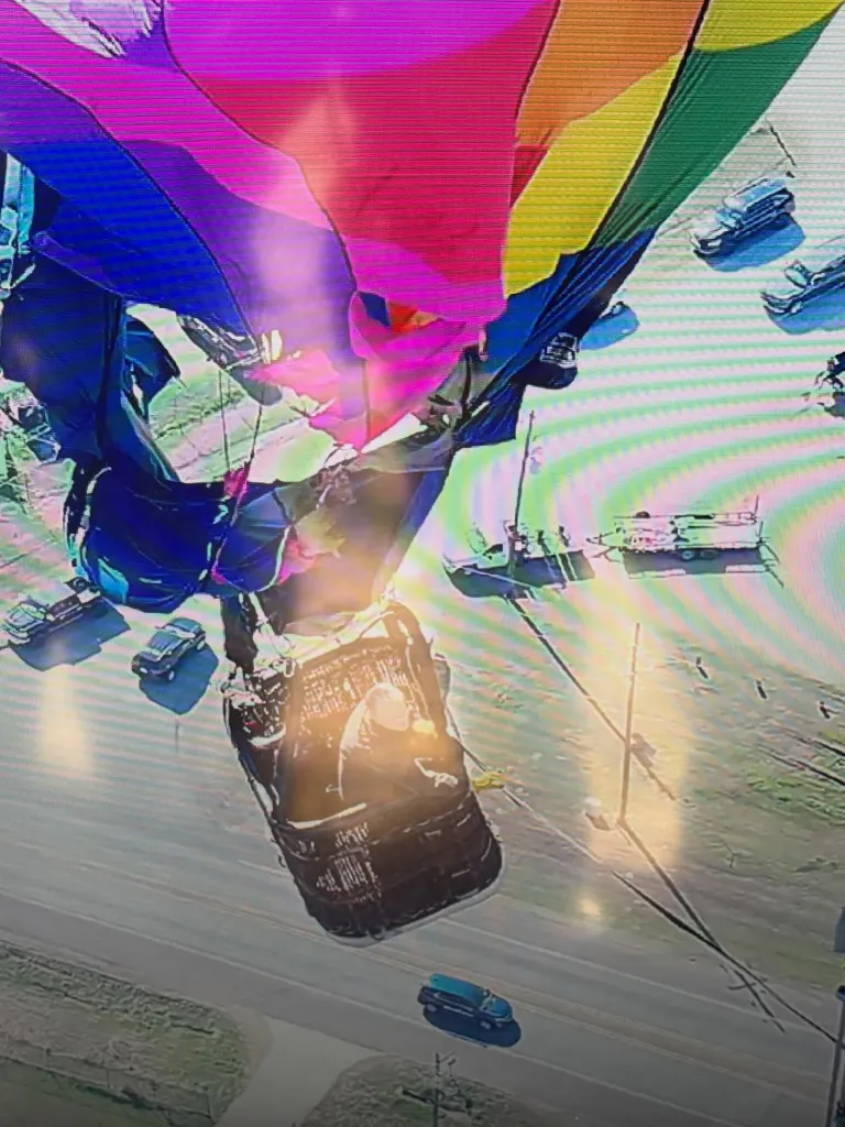 An aerial view shows a hot air balloon, with a rainbow-colored balloon and dark basket, hovering near a road with cars and trailers.