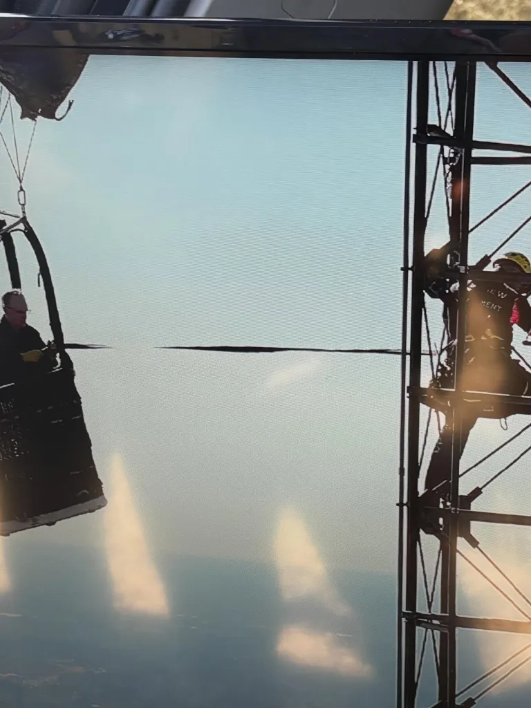 A hot air balloon pilot in a basket looking at a worker climbing a radio tower, with a wire connecting them.