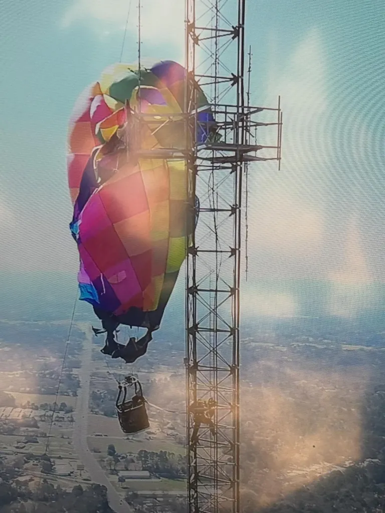 A colorful hot air balloon entangled with a tall communication tower, with a person climbing the tower and another in the basket.