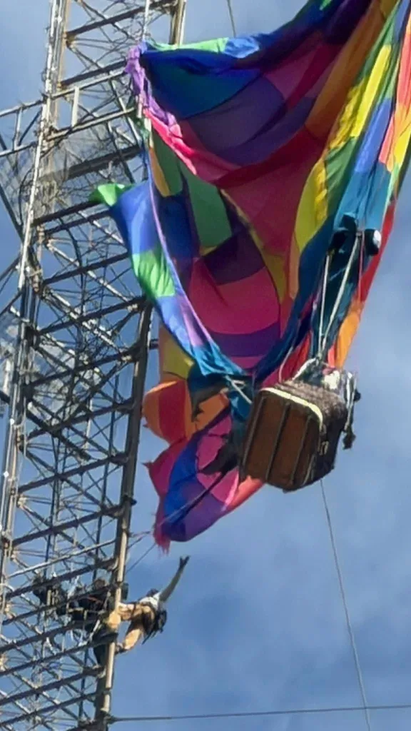 A person hanging from a broadcasting tower reaches out to a colorful parachute caught on the tower.