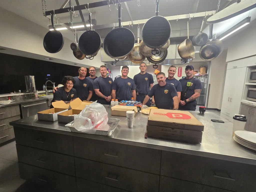 Ten firefighters in a kitchen with food and drinks on the counter.