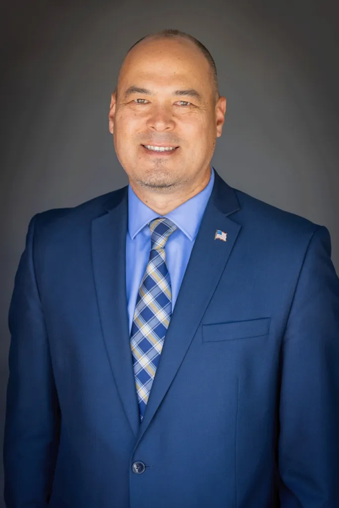 A man in a blue suit, blue shirt, and blue and yellow plaid tie. He has an American flag pin on his lapel.