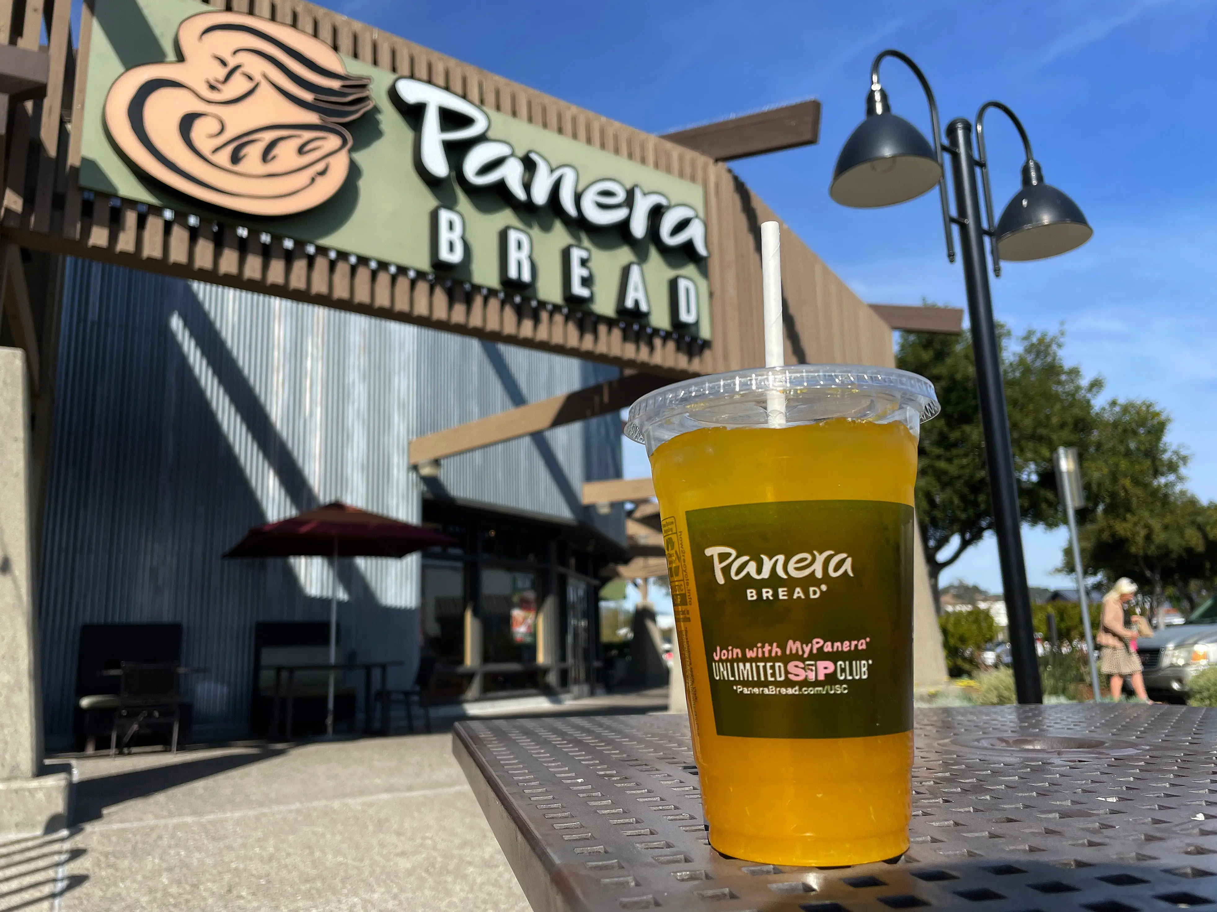 Panera Bread drink on a table outside in the foreground with Panera Bread restaurant in the background.