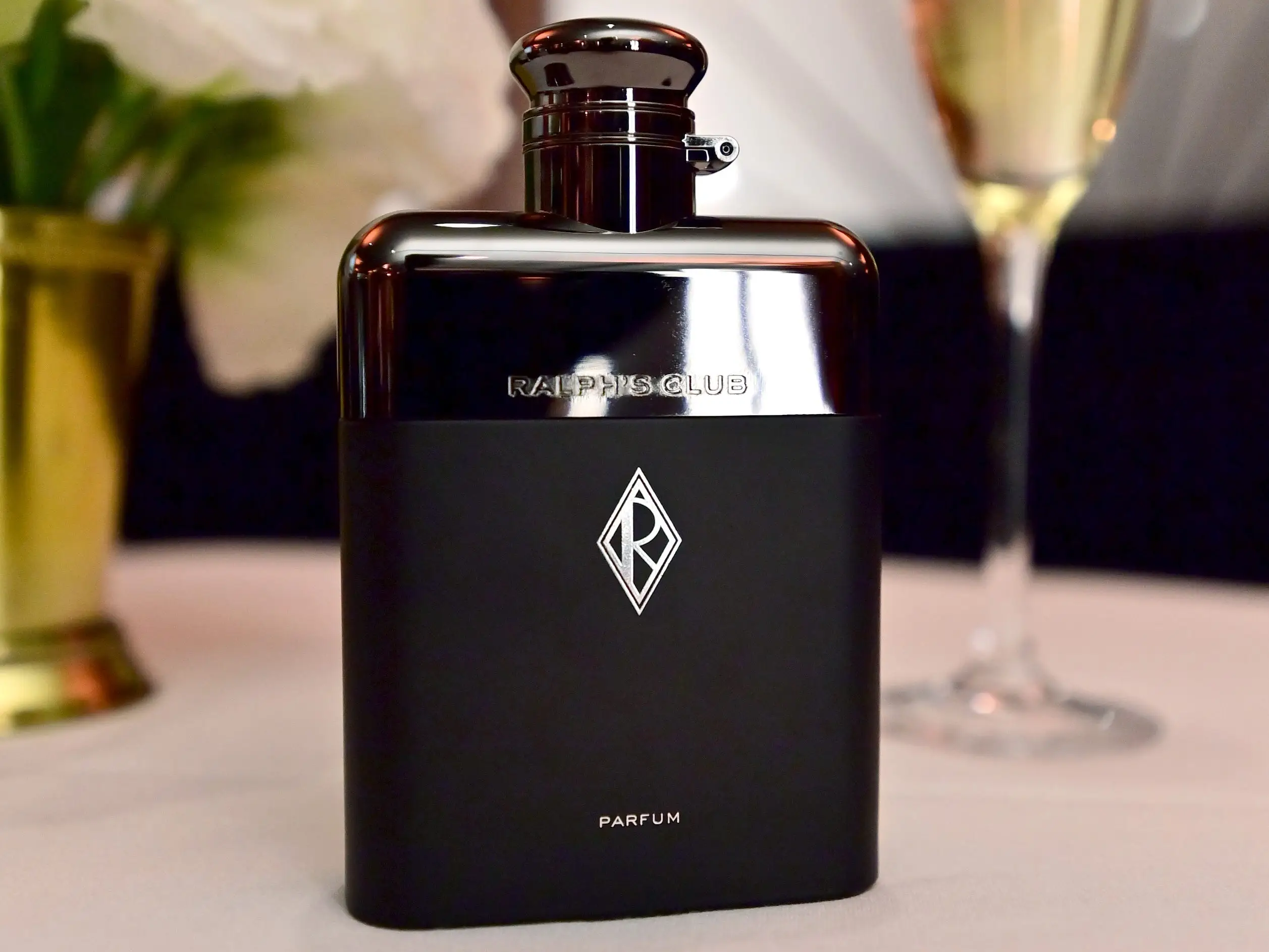 Bottle of Ralph's Club Parfum by Ralph Lauren with white flowers and a glass of sparkling wine in the background.