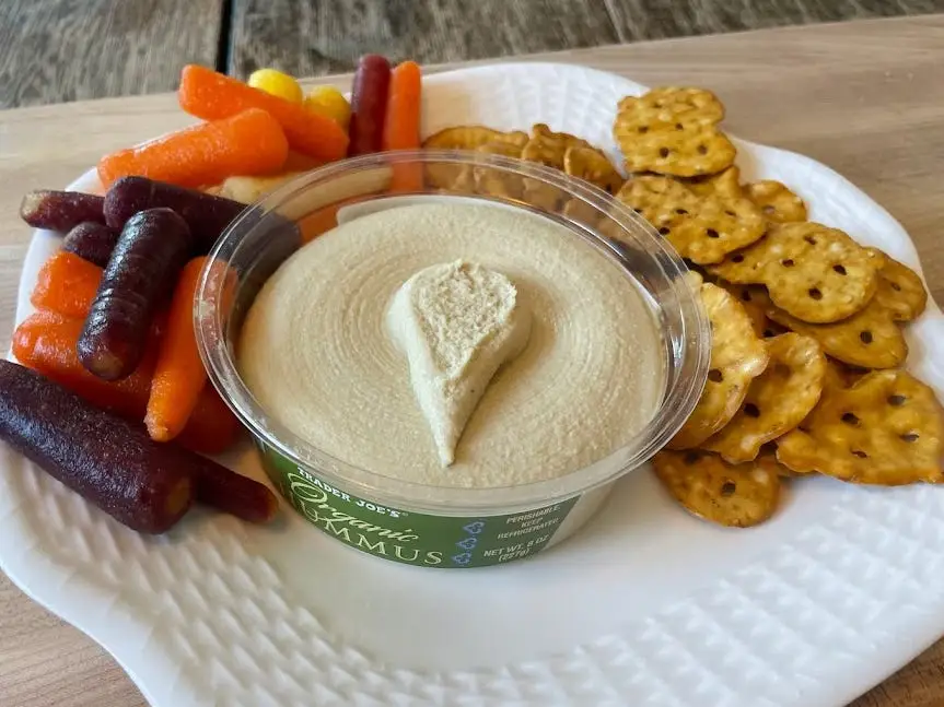 Container of Trader Joe's organic hummus, with a green label, on a plate with multicolored carrots and pretzel crisps