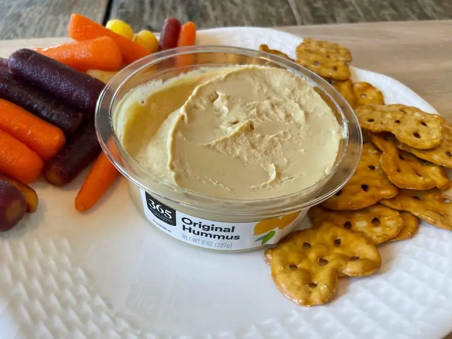 Container of 365 original hummus on a white plate with multicolored carrots and pretzel crisps