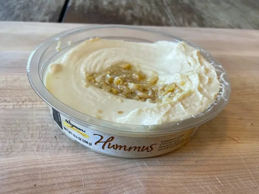 Container of Wegmans hummus with chickpeas on top of hummus on a wooden cutting board