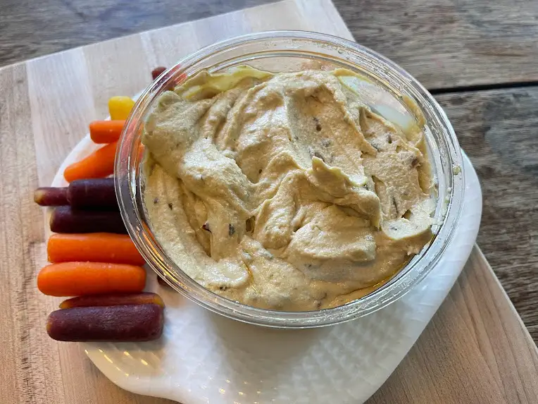 Container of stirred Kirkland Signature hummus with visible pieces of pine nuts in it. The container sits on a white plate with multicolored carrots