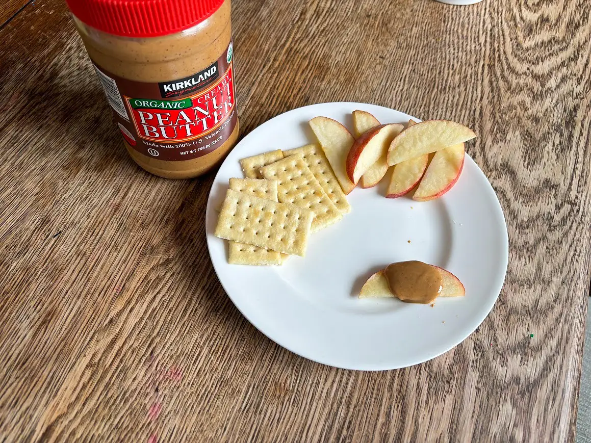A jar of peanut butter with a brown and red label sits next to a white plate of crackers and apple slices. Creamy-looking peanut butter drips down an apple slice on the plate