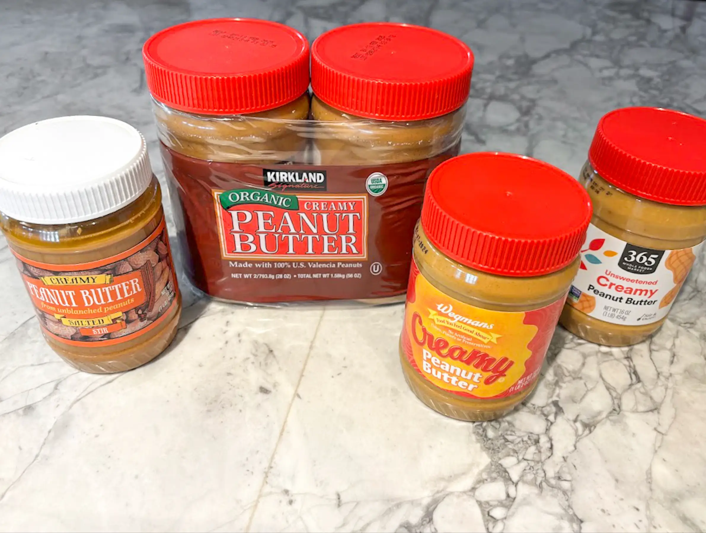 Jars of peanut butter on a white-marble counter. There is a jar of Trader Joe's peanut butter, a two-pack of Kirkland Signature peanut butter, a jar of Wegmans peanut butter, and a jar of 365 peanut butter