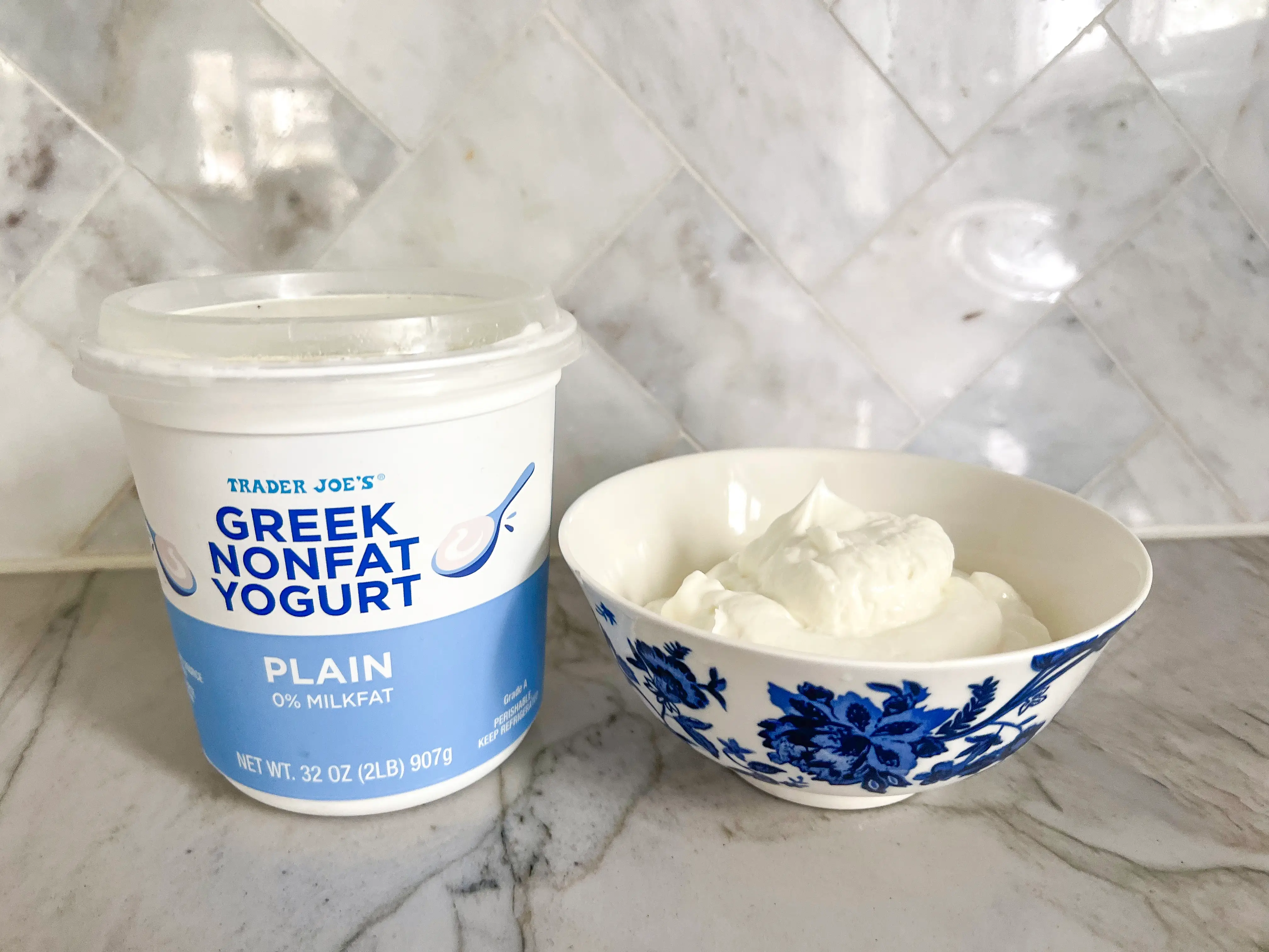 A white and light-blue container of Trader Joe's Greek nonfat yogurt with illustrations of spoons of yogurt on the container. A blue-and-white bowl filled with white yogurt sits next to the container