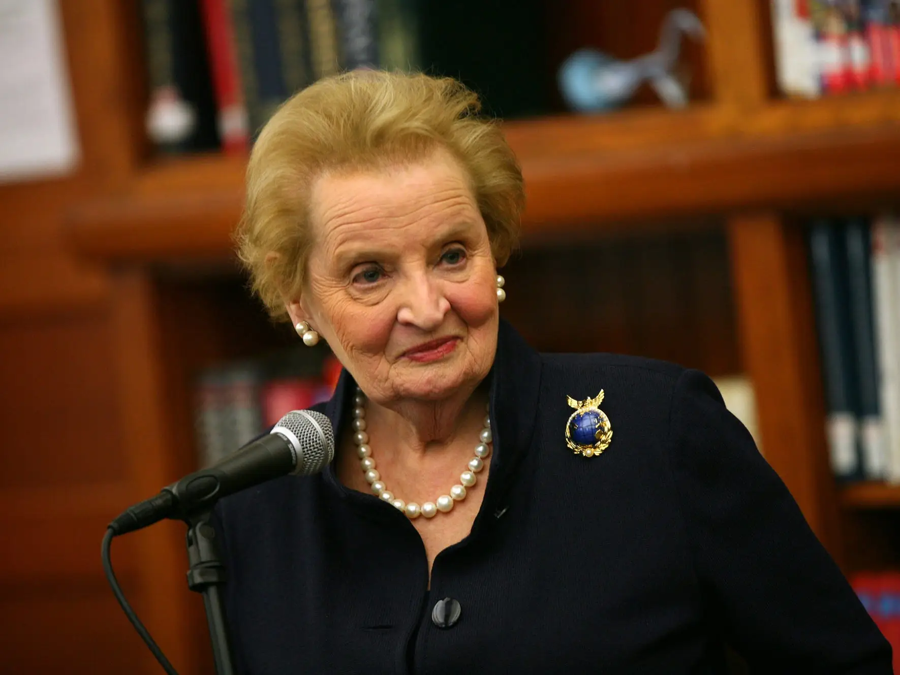 Madeleine Albright stands in front of a microphone in front of shelves of books in 2012
