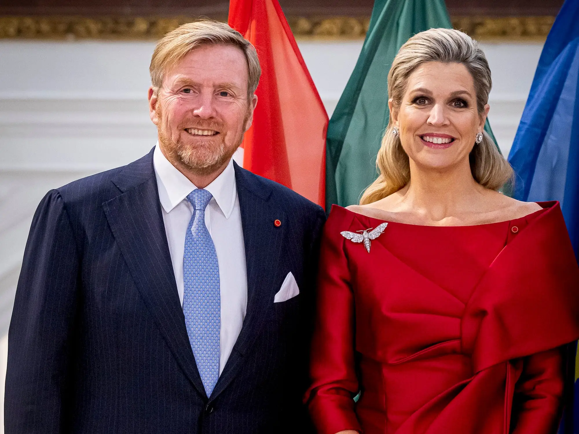 King Willem-Alexander in a suit and blue tie and Queen Maxima in a red dress with a sparkly dragonfly pin on her shoulder stand next to each other