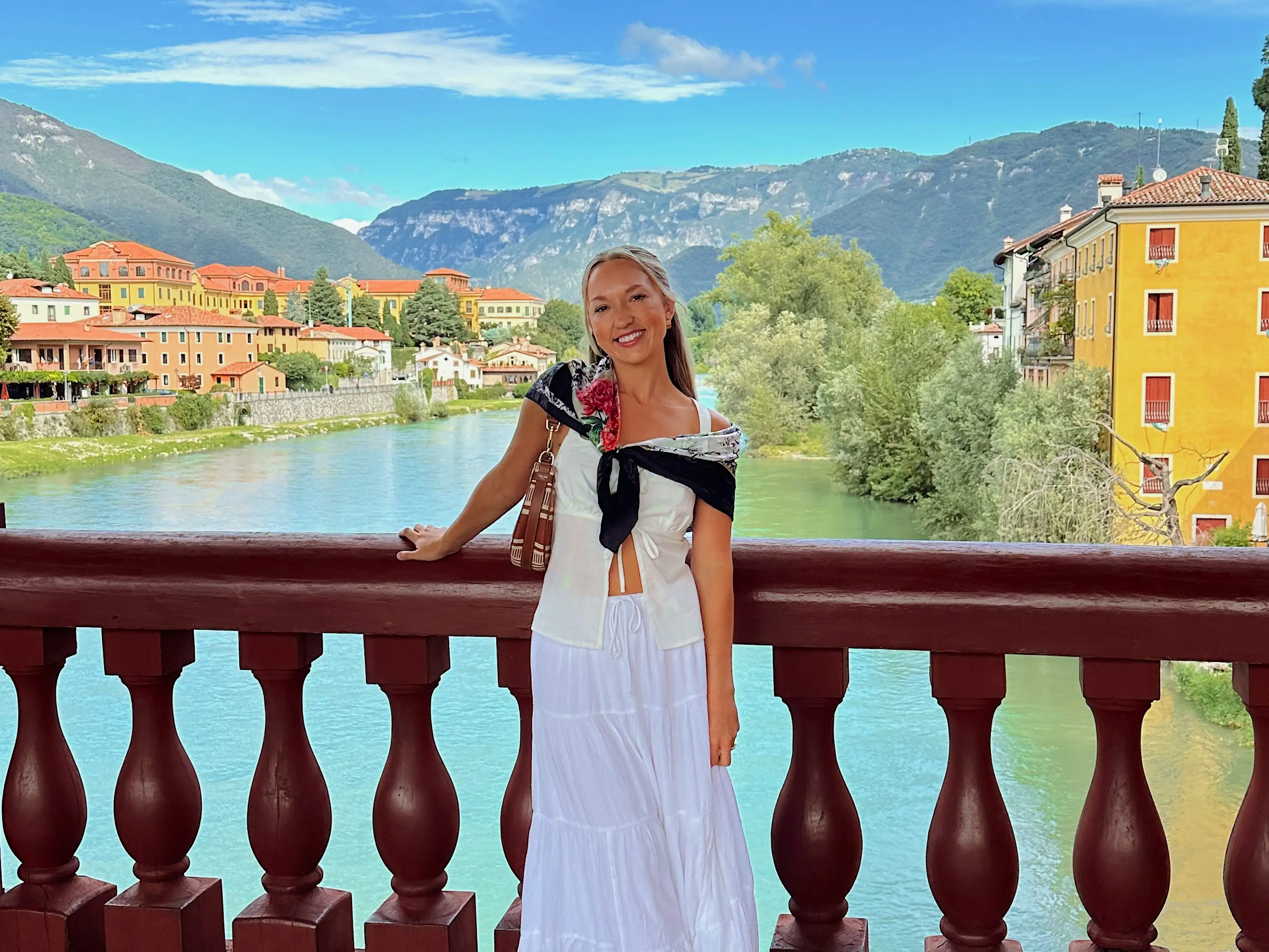 Chloe poses on a bridge in front of a river, yellow buildings with orange roofs, and mountains on a sunny day.
