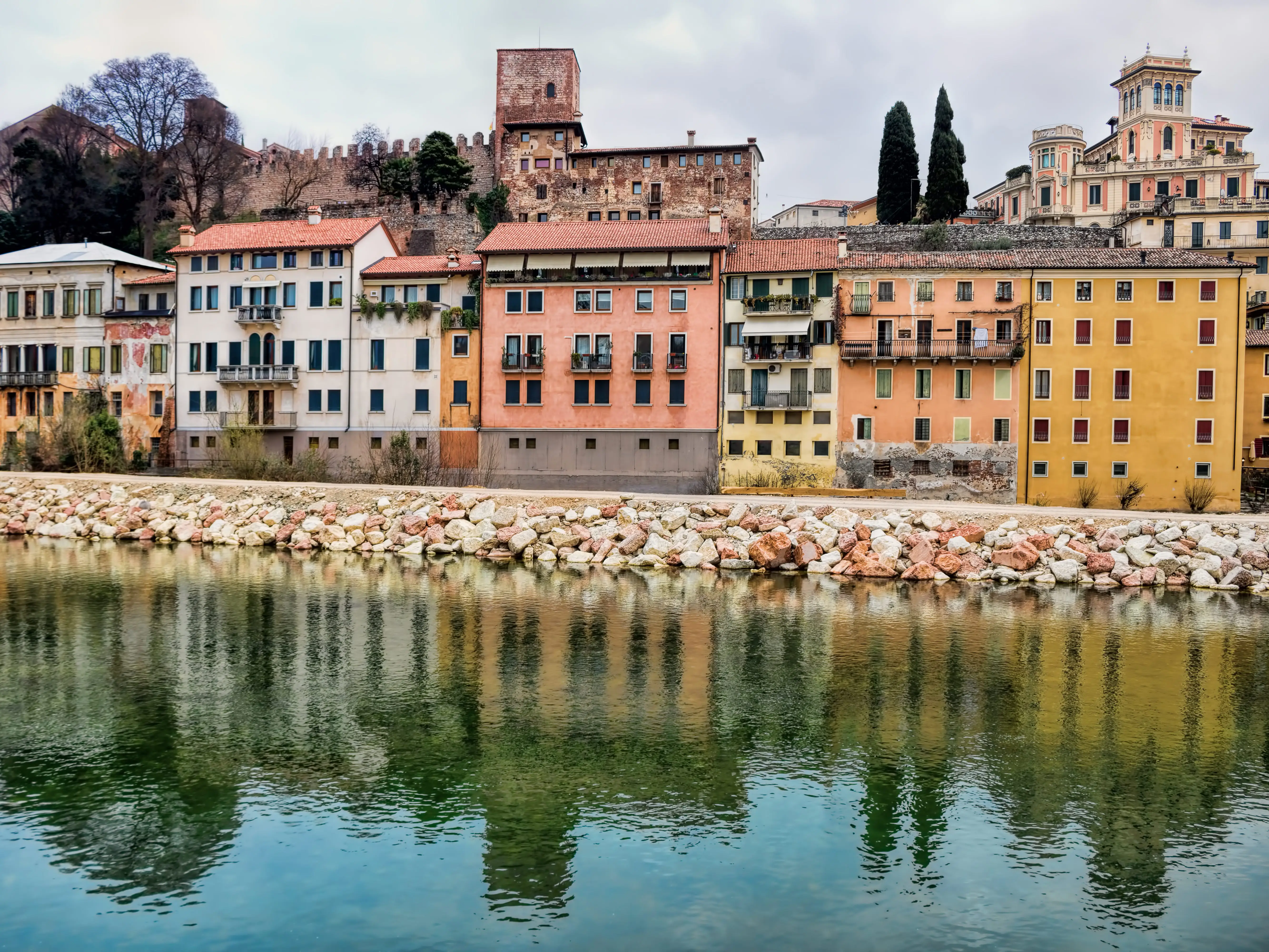 Old buildings and a dirt path line a river in Italy.
