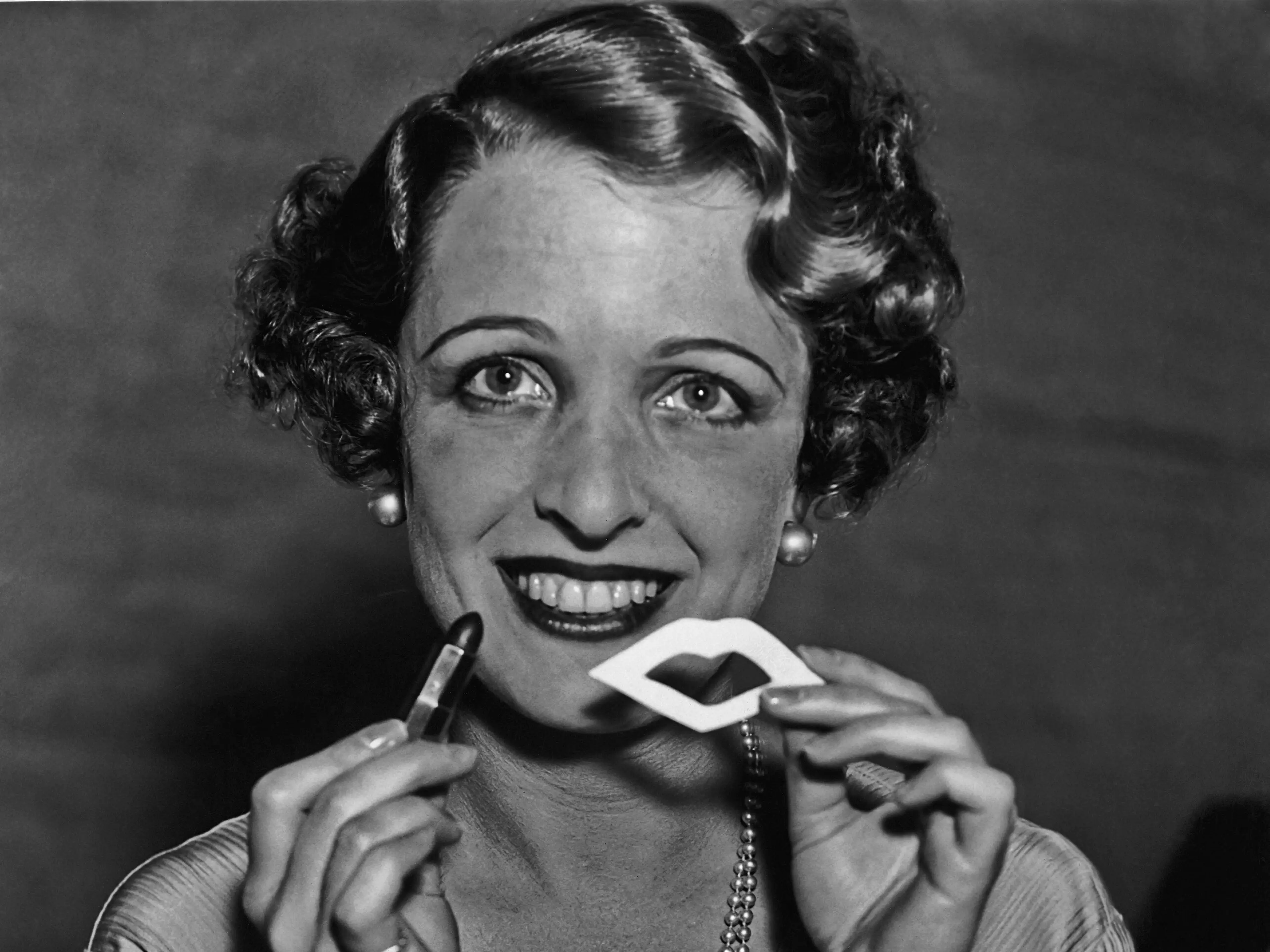 A woman smiled and held her lipstick and a stencil used to provide the perfect Cupid's Bow in 1925.