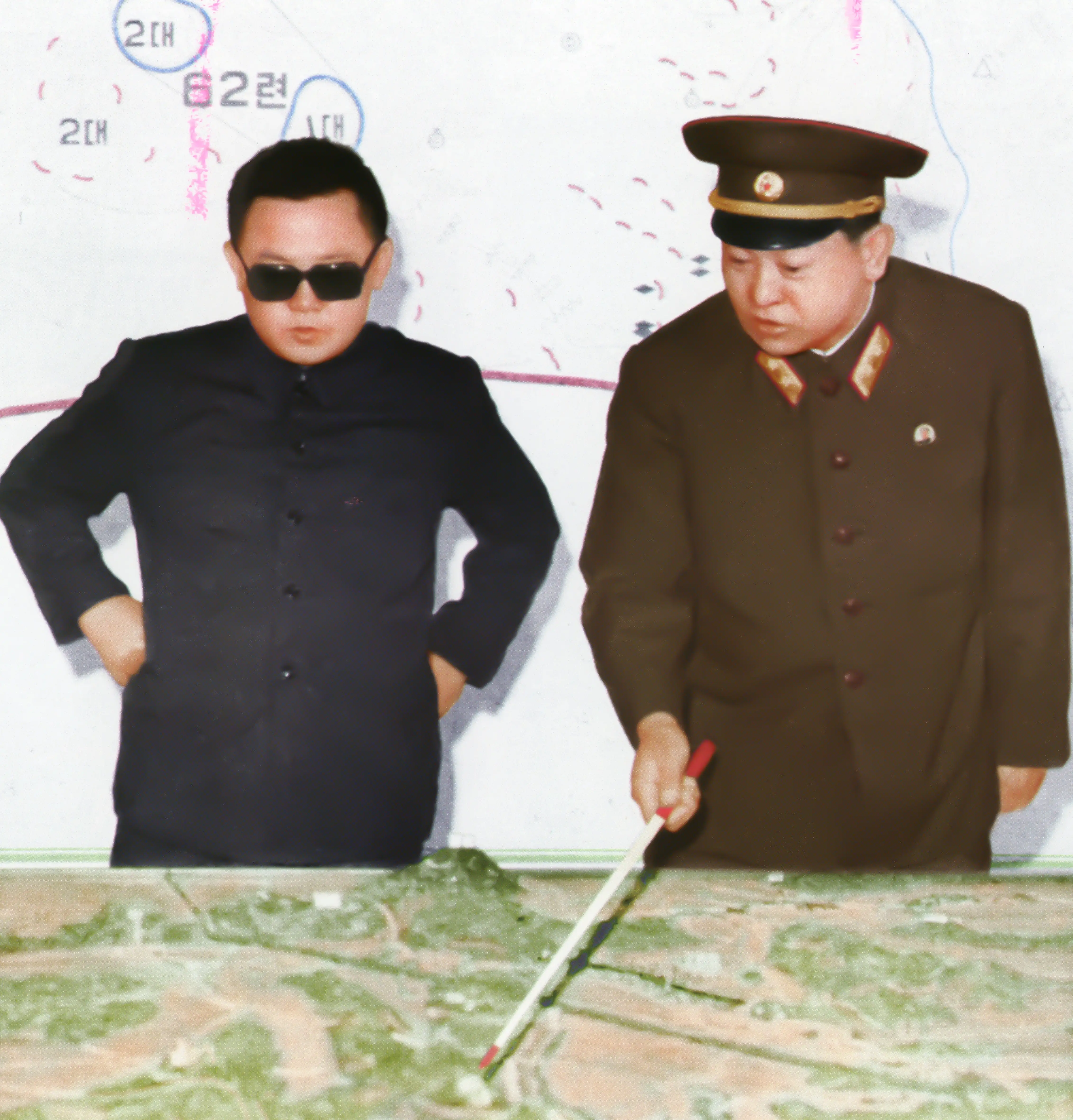 Kim Jong Il, wearing sunglasses and all black, and Kim Il Sung, in a military uniform and holding a red and white stick, look at a map