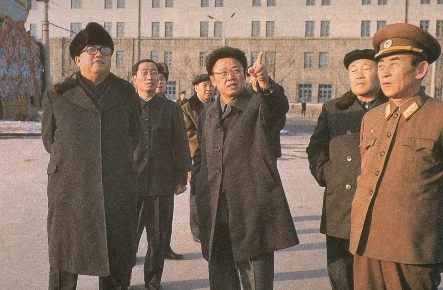 Kim Jong Il points standing next to his father who wears a coat and winter hat among others in uniforms and suits