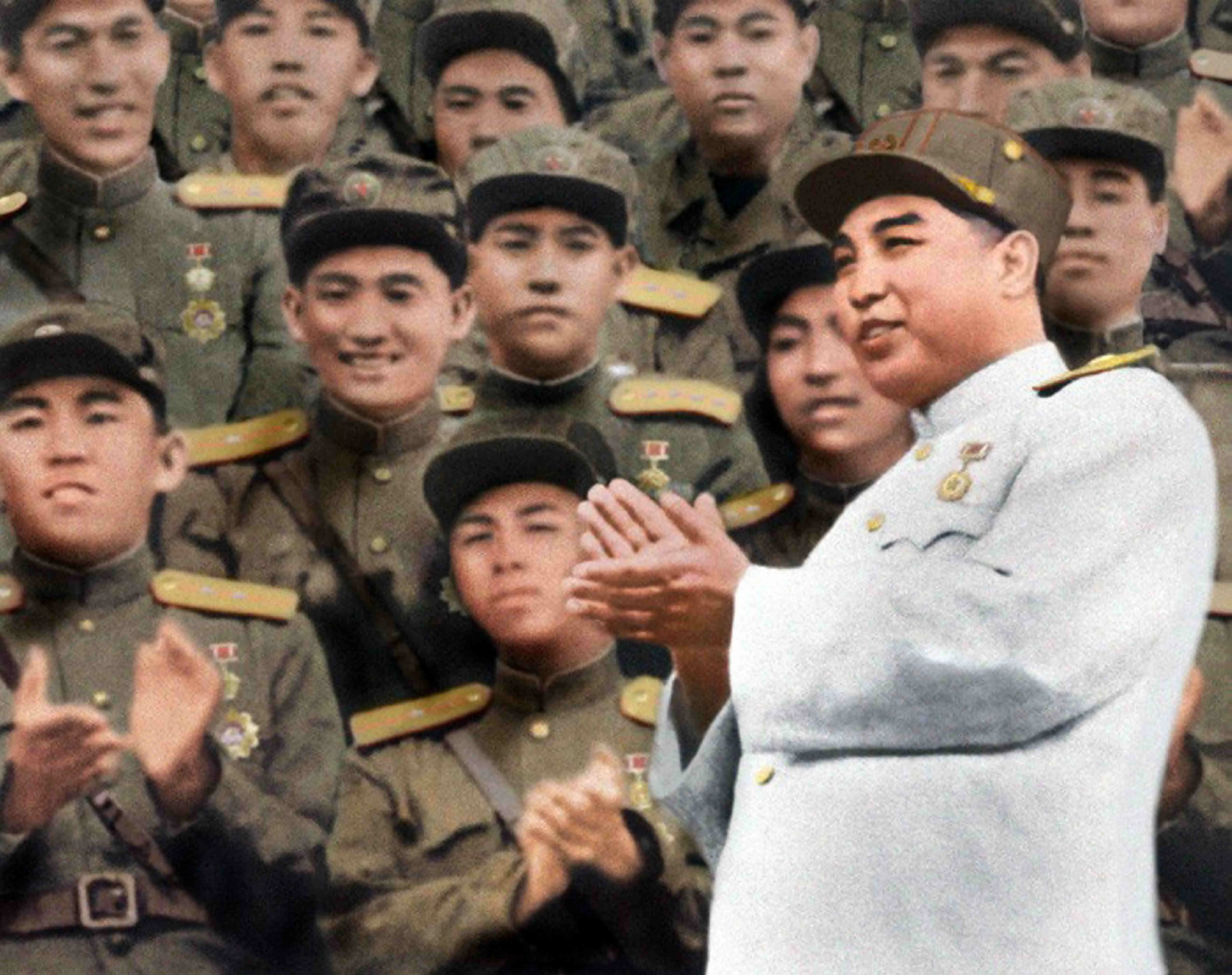 Kim Il Sung wears a gray military uniform and green hat in front of soldiers