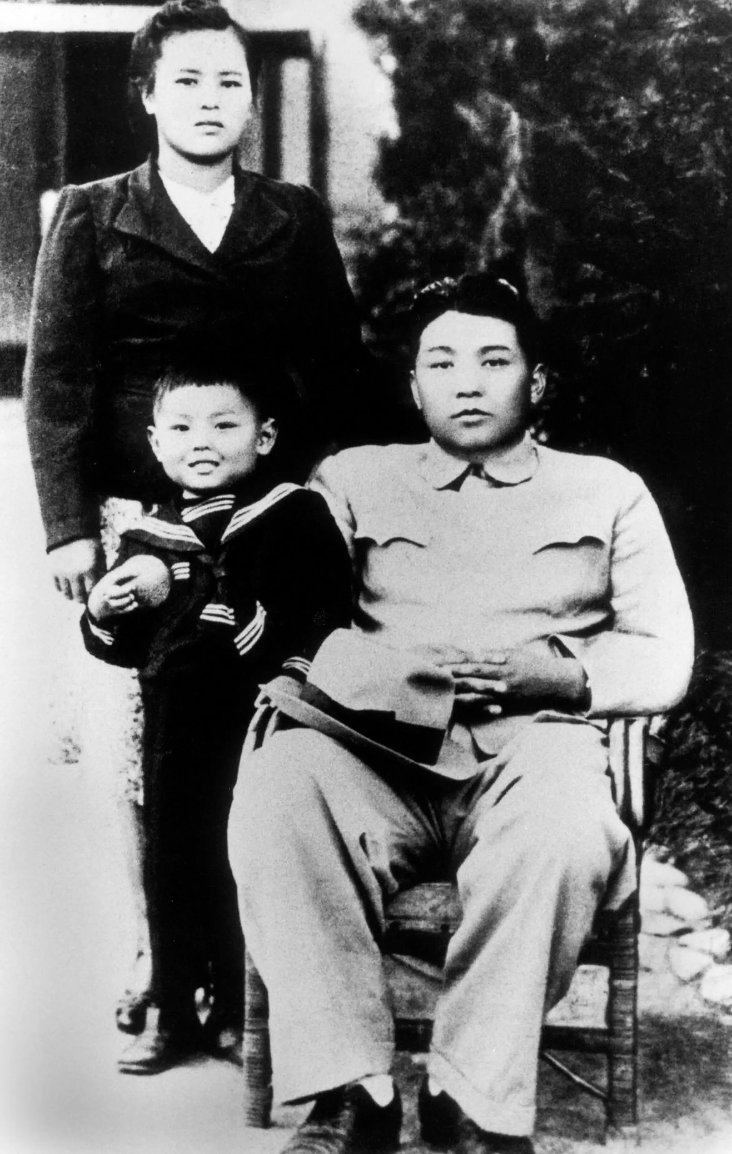 Kim Jong Il wears a sailor suit and stands in front of his mother while his father Kim Il Sung sits in a chair outdoors