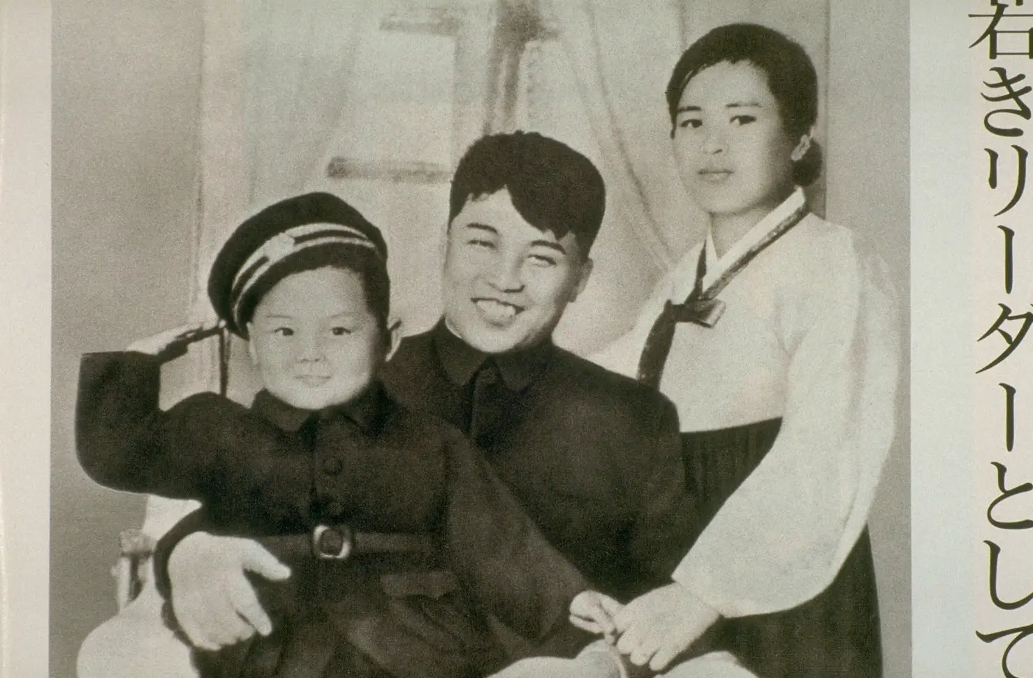 Young Kim Jong Il wears a military cap and salutes sitting in Kim Il Sung's lap and holding Kim Jong Suk's hand